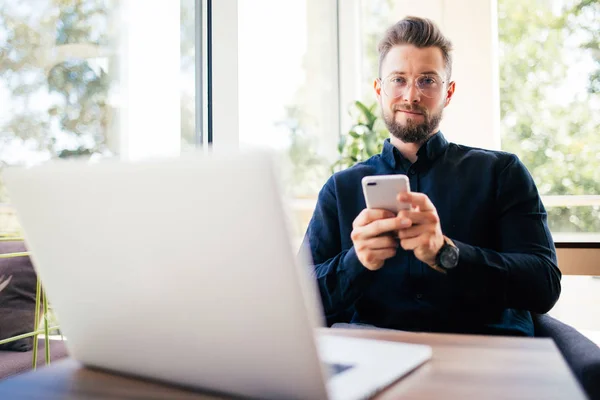 Joven hombre de negocios feliz sonriendo sentado en la oficina con el ordenador portátil mientras lee su teléfono inteligente. Retrato del hombre de negocios sonriente leyendo el mensaje con el teléfono inteligente en la oficina . — Foto de Stock