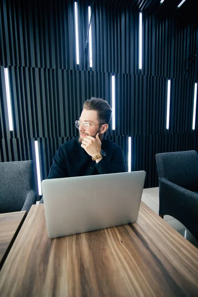 Atractivo hombre barbudo joven en traje mirando hacia otro lado mientras está sentado en el escritorio de la oficina con el ordenador portátil y otros artículos — Foto de Stock