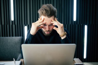 Ofiste oturan ve dizüstü bilgisayarda çalışan düşünce ve endişeli adam dalgın genç sakallı. Beyaz erkek ofisten çalışma.