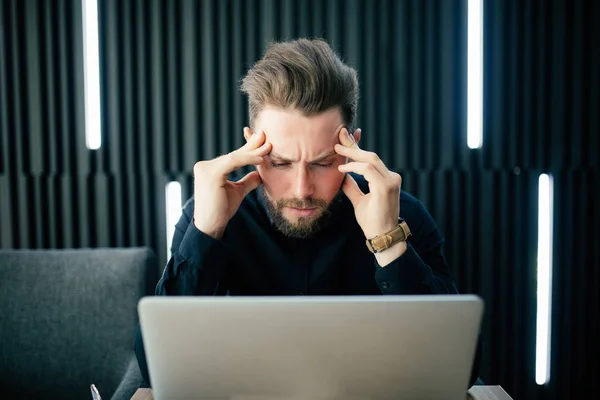 Nadenkend jonge bebaarde denken en bang man zittend op kantoor en werken op de laptop. Kaukasisch mannetje werken vanuit kantoor. — Stockfoto