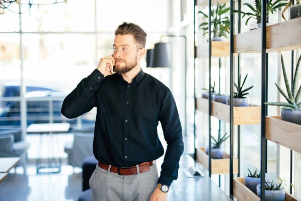 Mladý vousatý obchodní muž v úřadu mluvit na mobilním telefonu v moderní kanceláři — Stock fotografie