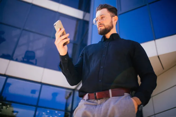 Portrét pohledný vousatý podnikatel při pohledu na mobilní telefon stojící poblíž kanceláře — Stock fotografie