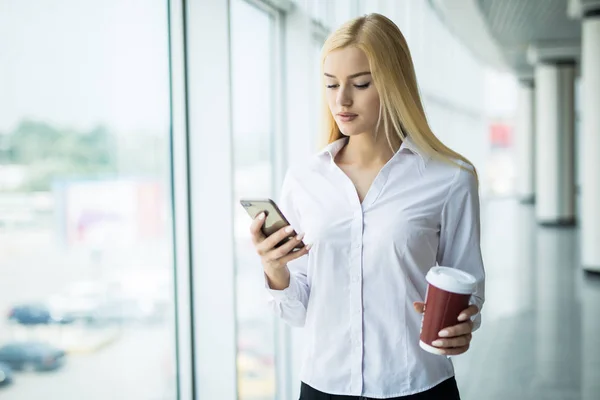Upptagen affärskvinna använda telefonen med kopp kaffe i andra hand nära stora panoramafönster på kontoret — Stockfoto