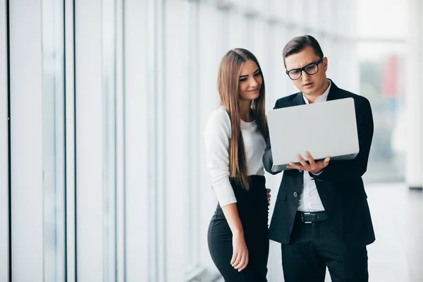 Empresario y mujer de negocios usando un portátil juntos mientras están de pie frente a las ventanas del edificio de oficinas en la oficina — Foto de Stock