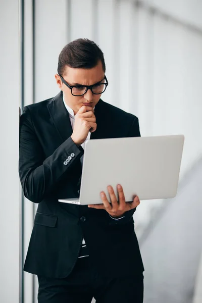Бізнесмен в окулярах і костюмі робочий ноутбук тримає руки біля панорамного вікна . — стокове фото