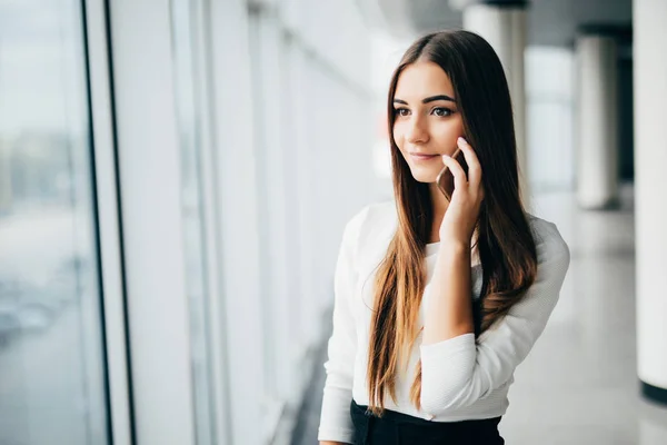Happy businesswoman standing in her office in a highrise building overlooking the cityscape. Businesswoman talking on mobile phone standing near window in office. — Stock Photo, Image
