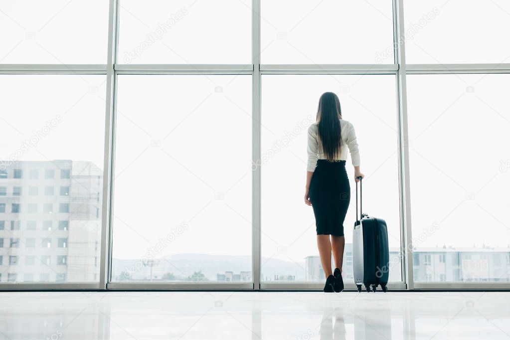Traveler businesswoman waiting for delayed flight at airport lounge standing with luggage watching tarmac at airport window. Woman at boarding gate before departure.