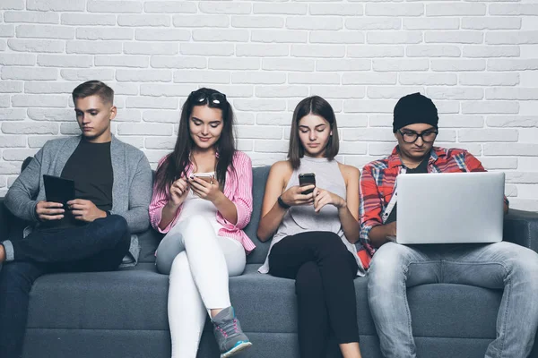 Hermosos jóvenes de diferentes nacionalidades están utilizando gadgets y sonriendo, de pie contra la pared de ladrillo blanco —  Fotos de Stock