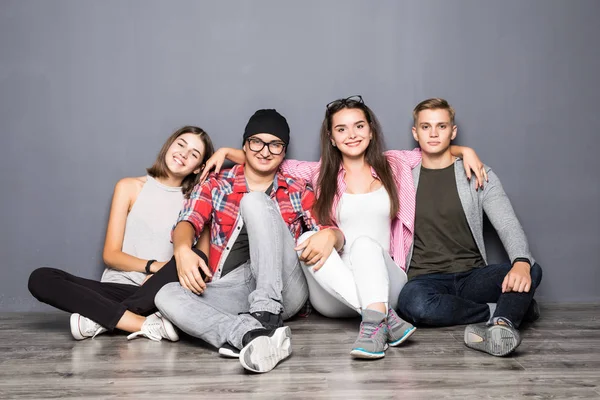 Schöne junge Freunde umarmen sich, blicken in die Kamera und lächeln, während sie auf dem Boden vor grauem Hintergrund sitzen — Stockfoto