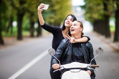 Güzel çift güneş gözlüklü bir akıllı telefon kullanarak ve bir scooter açık havada üzerinde otururken gülümseyen selfie yapıyor
