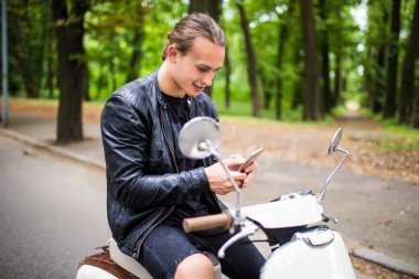 Scooter üzerinde oturan ve akıllı telefon kullanan genç yakışıklı adam