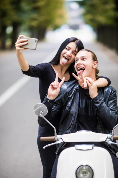 Красивая молодая любящая пара, сидящая вместе на скутере и делающая селфи на смартфоне, корча рожи — стоковое фото