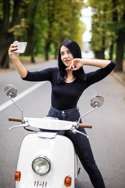 Açık selfie akıllı telefonunu alarak scooter üzerinde oturan genç ve güzel kadın portresi.