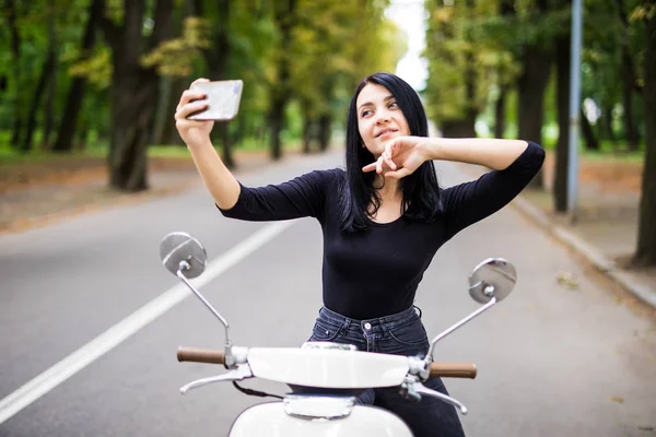Bir vintage scooter ve yapmak bir selfie telefon şehirde oturan genç kadın