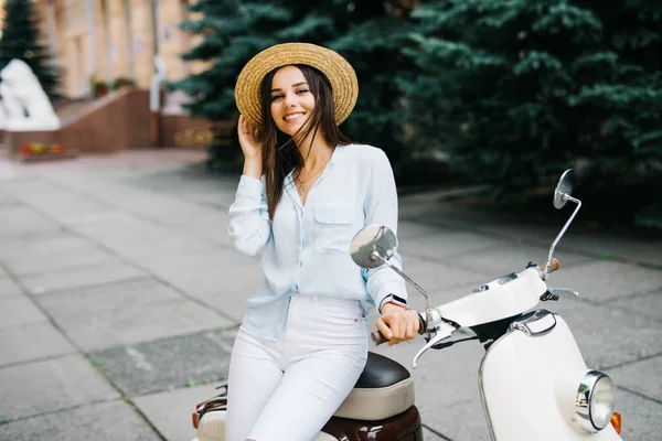 Feche a imagem de estilo de vida de mulher na moda jovem na roupa casual que se senta na scooter na rua. Vestindo camisa azul, calças brancas, óculos de sol na moda . — Fotografia de Stock