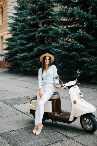 Feche a imagem de estilo de vida de mulher na moda jovem na roupa casual que se senta na scooter na rua. Vestindo camisa azul, calças brancas, óculos de sol na moda . — Fotografia de Stock