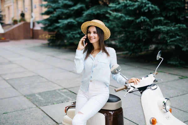 Güzel genç kadın rahat scooter üzerinde oturan ve telefonda konuşurken giyinmiş. Bisiklet üzerinde oturan ve hücresel smartphone kullanarak kız