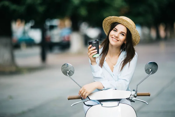 Jovem mulher na moda em roupa casual sentado em scooter e beber café na rua . — Fotografia de Stock