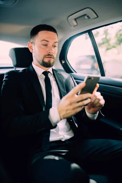 Ein Geschäftsmann, der mit dem Auto auf dem Rücksitz unterwegs ist, sendet eine Nachricht oder E-Mail und ruft an. der Mann in der Fahrer-Karriere für seine Dienstreisen. — Stockfoto