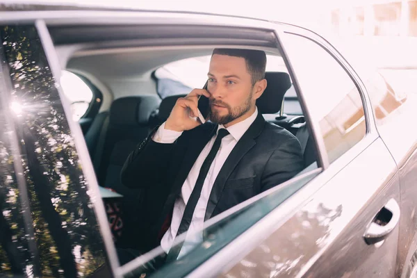 Joven hombre de negocios hablando por teléfono móvil y mirando hacia otro lado mientras está sentado en el asiento trasero de un coche — Foto de Stock