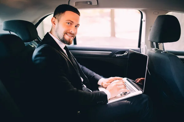 Bonito empresário barbudo trabalhando com laptop no banco de trás do carro — Fotografia de Stock