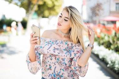 Genç kadın yürüyüş ve bir akıllı telefon sokakta bir güneşli yaz günde kullanarak.