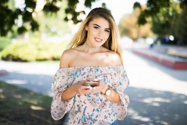 Genç kadın yürüyüş ve bir akıllı telefon sokakta bir güneşli yaz günde kullanarak.