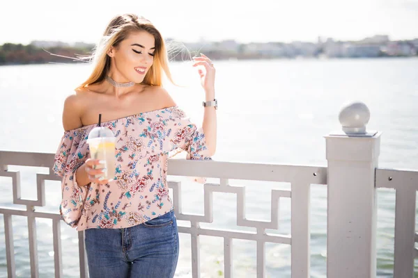 Menina sorrindo jovem sentado no dique do rio na cidade e beber limonada. Retrato de estilo de vida ao ar livre — Fotografia de Stock
