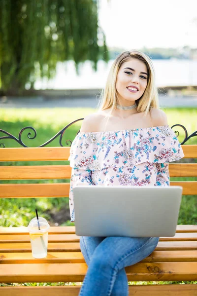 Молодая блондинка-студентка, сидя на скамейке, пользуется портативным ноутбуком. Молодая красивая девушка с помощью персонального ноутбука ищет информацию в Интернете — стоковое фото