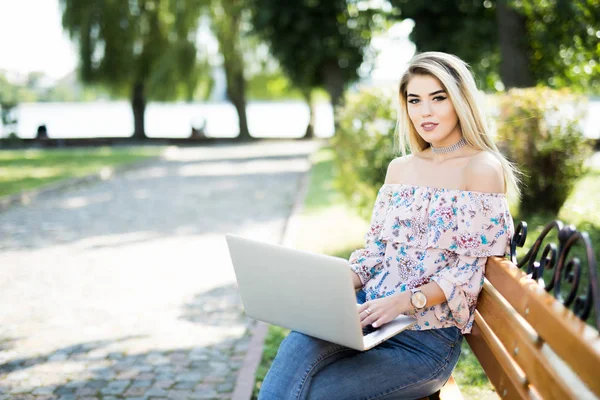 Блондинка с ноутбуком, расслабленно сидя на скамейке в летнем парке — стоковое фото