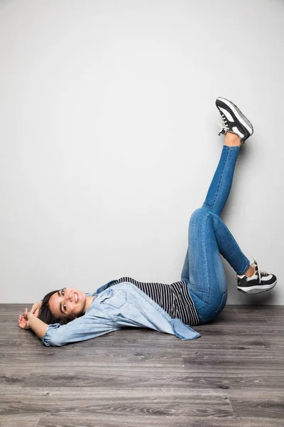 Szczęśliwy dzień kobieta leżącego na podłodze, z podniesionej nogi na szarym tle i patrząc na kamery — Zdjęcie stockowe