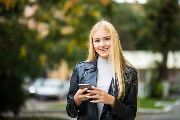Vacker ung kvinna tittar på smart telefon i city. unga leende kvinna som tittar på sin smarta telefon — Stockfoto