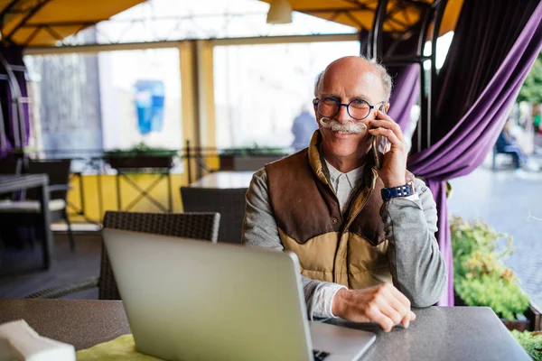 Portrait of senior man speaking by phone smiling happily while working with laptop in outdoor cafe lounge during coffee break