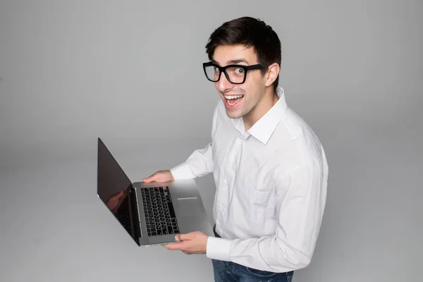 Guapo joven hombre de negocios en camisa y gafas está utilizando un ordenador portátil, mirando a la cámara y sonriendo, sobre fondo gris —  Fotos de Stock