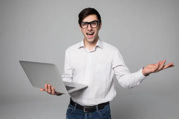Bonito jovem empresário de camisa e óculos está usando um laptop, olhando para a câmera e sorrindo, em fundo cinza — Fotografia de Stock