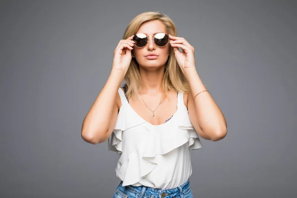 Mladá atraktivní dáma v džínách a sluneční brýle pózuje v studio na šedém pozadí není izolovaná, plavovláska bokovky letní oblečení koncept, Foto s místem pro text — Stock fotografie