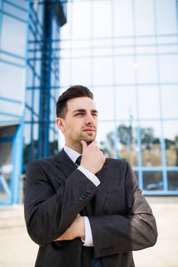 Takım elbiseli çeneme elini tutarak ve uzak ofis binası arka planda ile ayakta açık havada süre seyir emin düşünceli işadamı