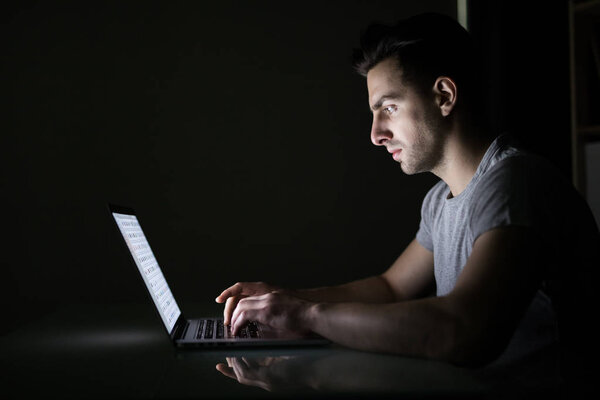 Молодой человек читает новости на ноутбуке, лежащем в темной комнате. Выглядит сосредоточенным
