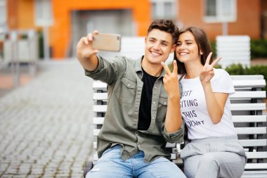 Happy young couple hugging and laughing outdoors on the bench with victory sign clipart