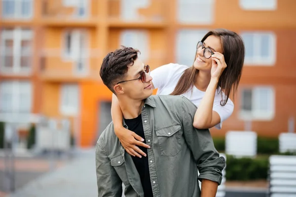 Bela jovem casal em óculos de sol olhando um para o outro e sorrindo enquanto de pé ao ar livre. Menina piggyback — Fotografia de Stock