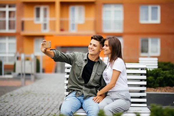 Pareja joven de compras. Hermosa pareja joven sentada en el banco y tomando una selfie con un teléfono móvil . — Foto de Stock