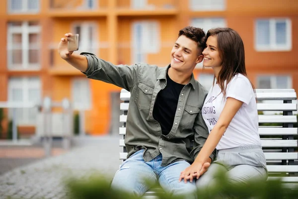 Pareja joven de compras. Hermosa pareja joven sentada en el banco y tomando una selfie con un teléfono móvil . — Foto de Stock