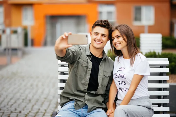 Hermosa pareja joven sentada en el banco en el centro de la ciudad y tomando una selfie con una tableta. Concepto de estilo de vida — Foto de Stock