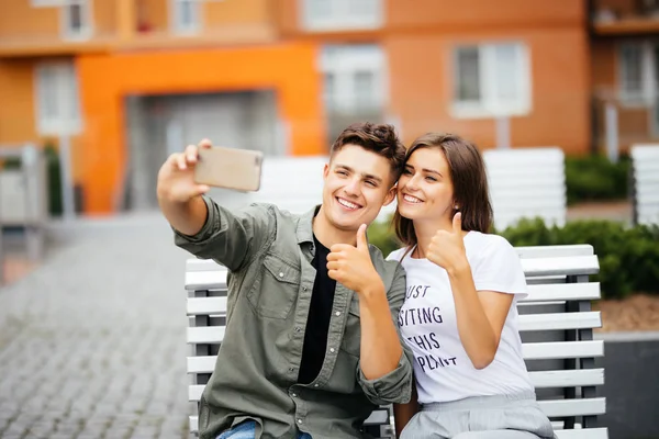 Hermosa pareja joven sentada en el banco en el centro de la ciudad y tomando una selfie con los pulgares hacia arriba. Amor y ternura. Concepto de estilo de vida — Foto de Stock