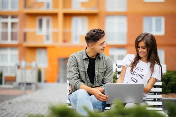 Gelukkige jonge paar laptopcomputer zittend op een bankje in de stad buiten gebruikt. Twee geliefden plezier tijd samen om op te letten sociaal netwerk — Stockfoto