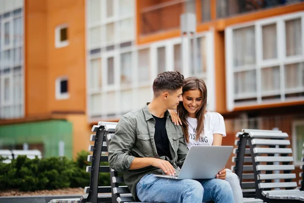 Feliz jovem casal usando laptop enquanto sentado no banco no parque e sorrindo uns aos outros — Fotografia de Stock