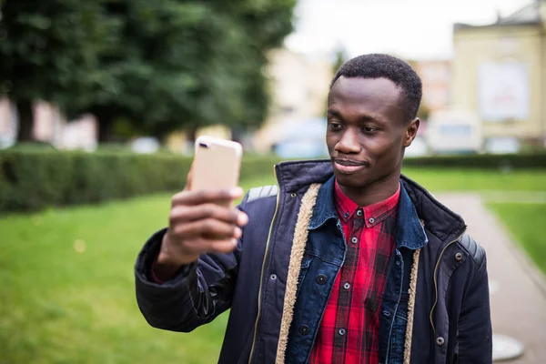 Foto seorang pria gembira african memegang ponselnya di tangan dan membuat selfie luar ruangan . — Stok Foto