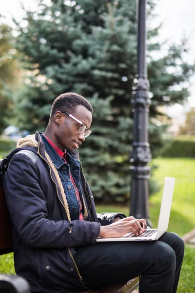 Jonge Afrikaanse man met laptop zittend op een houten bankje en typen. — Stockfoto