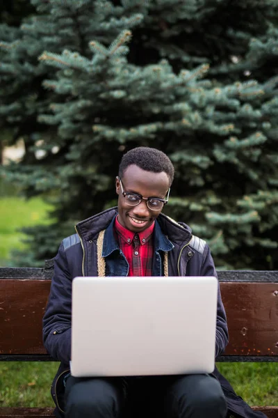 Foto eines jungen, fröhlichen Mannes aus Afrika, der auf einer Bank sitzt und einen Laptop benutzt. Blick in die Kamera. — Stockfoto