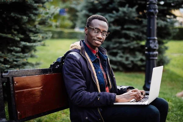 Joven africano con portátil sentado en un banco de madera y mecanografía . — Foto de Stock